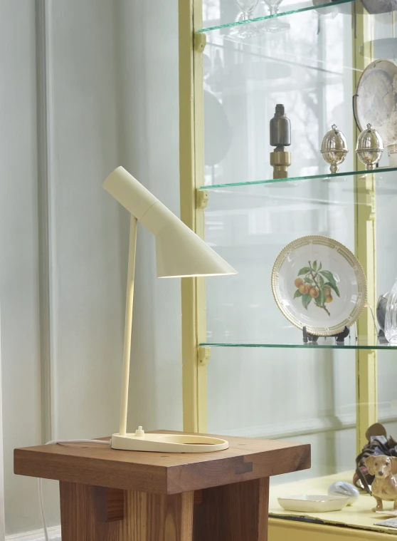 クリーム色のAJ Table Lampが木のテーブルに置かれ、ガラス製品や磁器の並ぶ飾り棚のそばにある。 © 2026 Louis Poulsen.