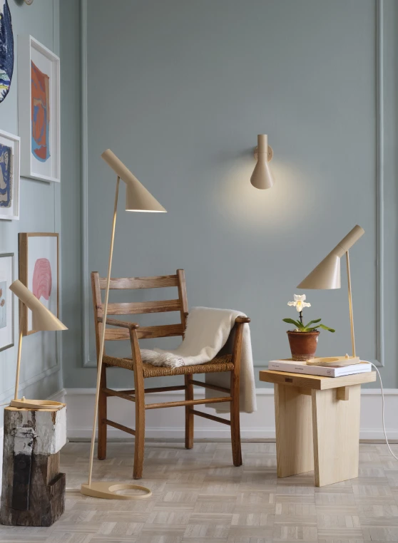 サンドカラーのAJ Floor Lamp、AJ Wall Lamp、AJ Table Lampが、落ち着いた室内の木製椅子を照らしています。 © 2026 Louis Poulsen.