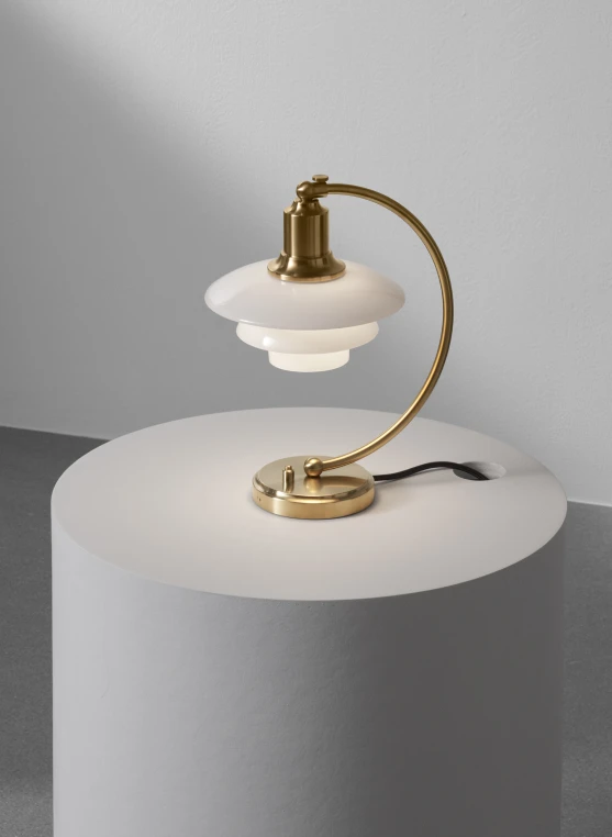 PH 2/2 Luna bordlampe i messing med hvidt opalglas står på en rund sokkel mod en lys væg. © 2026 Louis Poulsen.