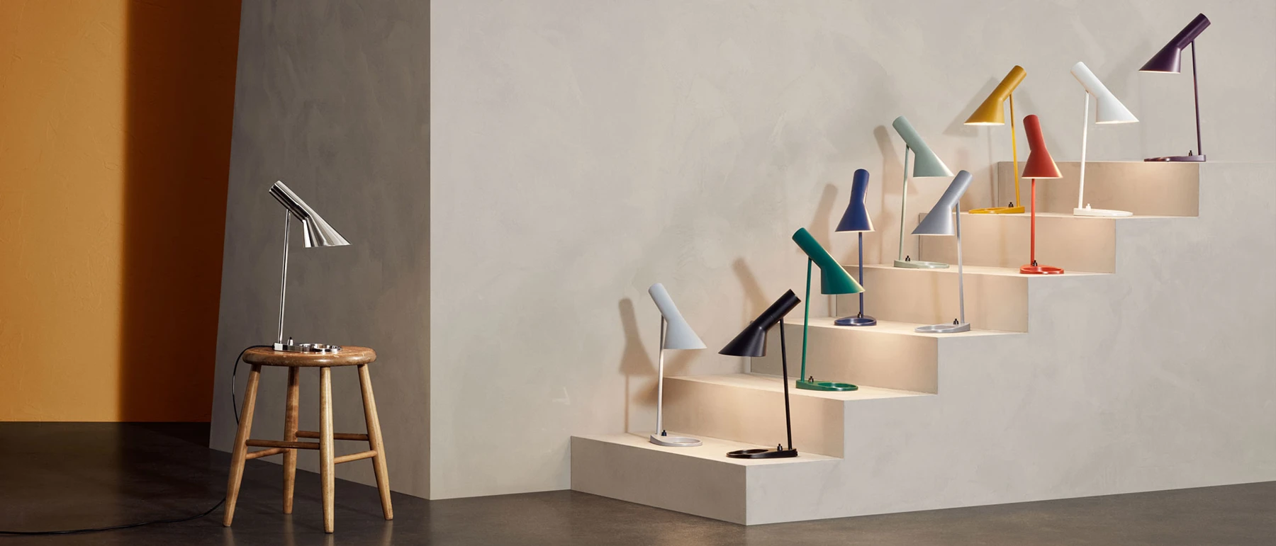 AJ Table Lamps in assorted colours line a stepped display, with a chrome version on a wooden stool beside them. © 2026 Louis Poulsen.