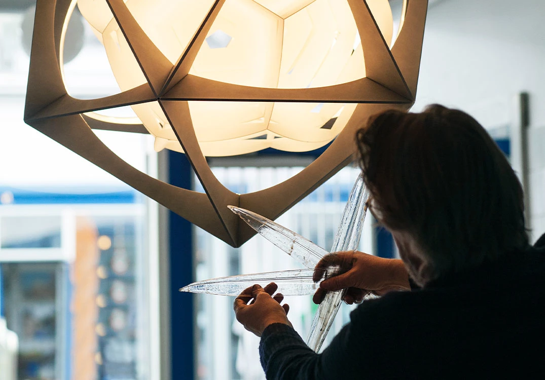 En oplyst OE Quasi Light hænger over en person, der arbejder med klare glaselementer i et lyst rum. © 2026 Louis Poulsen.